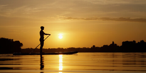 Silhouette of woman standing on SUP board and paddling at sunset, light waves on water. Pan shot doing sports in beautiful landscape. Healthy lifestyle. Concept of fitness