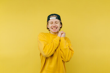 Joyful young man in a yellow sweatshirt stands on a yellow background with a happy face and closed eyes.