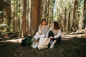 Female friends relaxing in the woods outdoors. Two beautiful women tourists are sitting in a mountain forest and relaxing, talking with smiles on their faces.