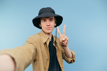 Cute young man in panama taking selfie on smartphone and showing peace gesture on blue background.