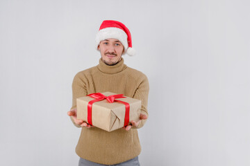 A young smiling Caucasian man wearing Santa Claus hat holding out gift and looking at camera on white isolated background