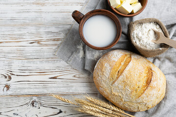 Fresh white bread with a crisp crust on a light napkin. A mug with milk, butter in a clay bowl and flour in a canvas bag in the background. Top view