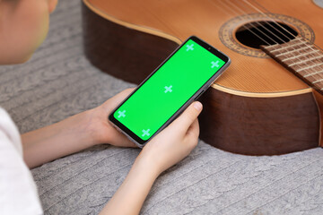 Selective focus composition with boy holding the smartphone with green screen on a blurred background with guitar. Mockup for mobile music apps. Guitar lesson online. Online music education - Powered by Adobe