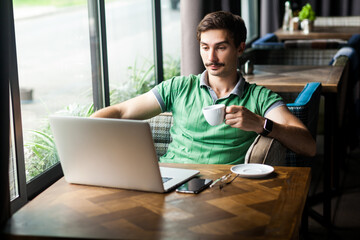 Portrait of relaxed calm handsome businessman wearing green T-shirt, enjoying hot coffee while working online on laptop, having break during work. Indoor shot near big window, cafe background.