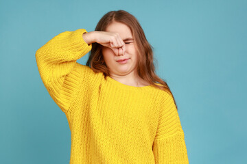 Little girl holding breath, pinching her nose and expressing disgust to stink, intolerable odor, wearing yellow casual style sweater. Indoor studio shot isolated on blue background.