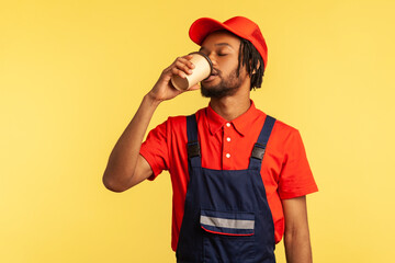 Portrait of bearded worker wearing red T-shirt and blue overalls, drinking coffee from disposable paper cup, likes take away beverage while working. Indoor studio shot isolated on yellow background.