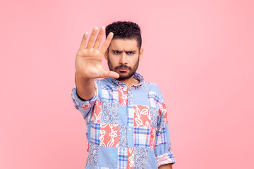Strict brunette bearded man showing stop sign at camera, wearing casual style blue suit standing with frowning face and bossy expression. Indoor studio shot isolated on pink background.
