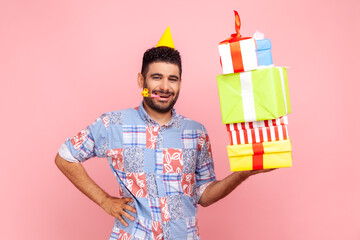 Happy positive man with beard wearing stack of presents boxes, blowing party horn, keeping hand on hip, looking at camera with satisfied expression. Indoor studio shot isolated on pink background.