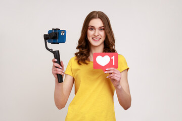 Smiling teenage girl shoots herself on electronic stabilizer for video shooting on phone, broadcasting livestream, showing like icon. Indoor studio shot isolated on gray background.