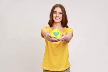 Smiling confident teenager girl in yellow casual style T-shirt holding paper house in hand, real estate agency, accommodation, buying own house. Indoor studio shot isolated on gray background.
