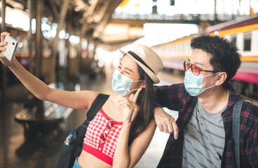 Asian backpack traveler people with camera standing at train station platform and waiting train arrivel, summer .trip in summer with face mask for protection by infection from Coronavirus, Covid-19