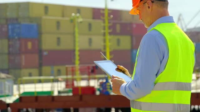 Cargo ship loading in dock. Mature general manager in uniform vest with tablet plans freight delivering in dockyard on sunny day close view