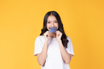 Portrait of a happy young asian woman wearing white shirt holding bank card, credit card isolated on yellow background. Business online shopping concept.