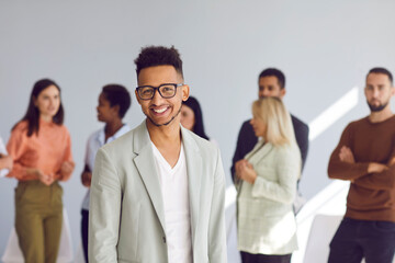 Obraz premium Portrait of positive male employee standing on blurred background of his colleagues. Friendly handsome dark-skinned office worker or businessman working in modern business center. Success concept.