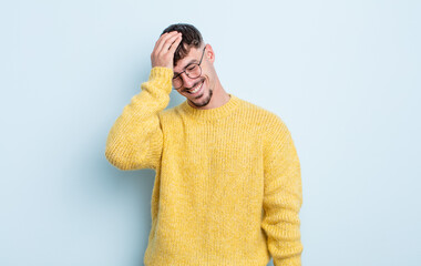 young handsome man laughing and slapping forehead like saying d’oh! I forgot or that was a stupid...