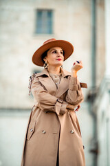 Outdoor fashion portrait of young elegant fashionable brunette woman, model in stylish hat, choker and light raincoat posing at sunset in European city.