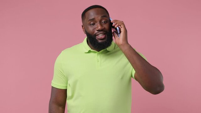Happy calm young bearded african american man 20s wears green t-shirt hold use talk on mobile cell phone conducting pleasant conversation isolated on plain pastel light pink background studio portrait
