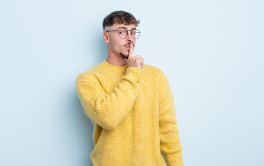 young handsome man asking for silence and quiet, gesturing with finger in front of mouth, saying...