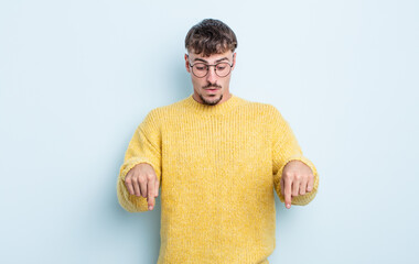 young handsome man with open mouth pointing downwards with both hands, looking shocked, amazed and...