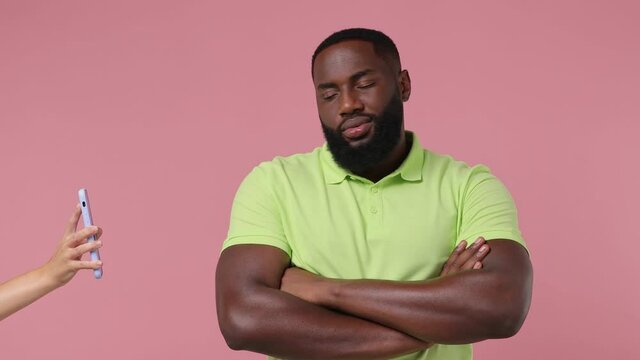 Surprised fun young bearded african american man 20s wears green t-shirt stand with crossed hands aside hand hold mobile cell phone show isolated on plain pastel light pink background studio portrait