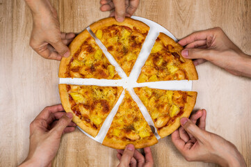 Top view of hands taking slices of delicious and crispy hawaiian pizza. Group of hungry friends sharing delicious lunch on wooden table.