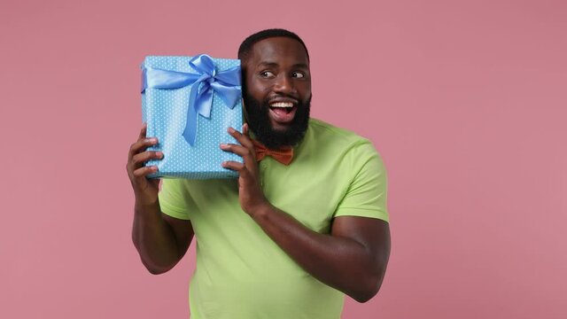 Joyful young african american man 20s wears green t-shirt bow tie hold shake try to guess what`s inside blue present box with gift ribbon isolated on plain pastel light pink background studio portrait