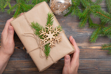 women's hands with a Christmas gift in craft paper with a fir branch, a ball, a snowflake on a brown wooden background