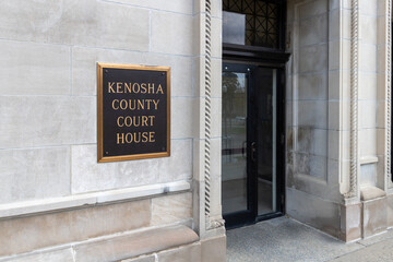 Exterior entry sign of the Kenosha County Court House.  Simple gold lettering on black background mounted on white marble wall.  Glass entry door visible with reflection. 