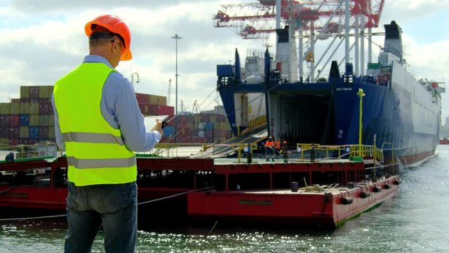 Marine port cargo industry. Safety manager holds tablet computer and portable radio while controls workers team near open ship hold in dock