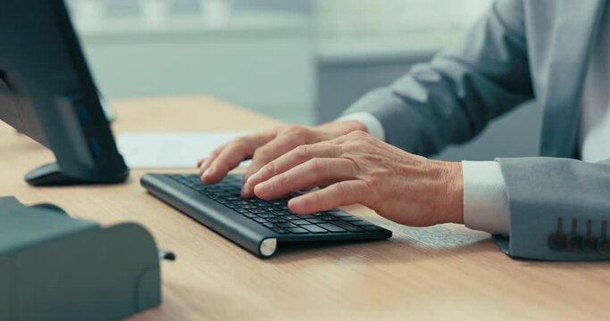Close-up of older man's hands on black computer keyboard, businessman in gray shirt and jacket, mature guy taps fingers on buttons, fills out company documents, boss behind desk