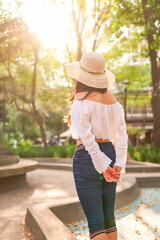 young latin tourist woman walking through a park full of trees at sunset, wearing a hat, blue pants, white blouse and a handbag, model not recognizable, with her back turned, vertical picture
