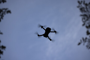 Silhouette of a small quadrocopter - drone against a background of blue sky and branches.