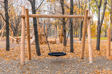Children's wooden playground in the autumn park. Wooden swing in the foreground. Logs, chains and ropes. Yellow fallen leaves