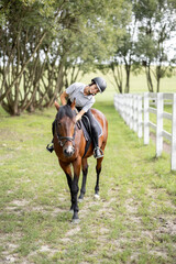 Female horseman riding brown Thoroughbred horse in green meadow in countryside. Concept of rural resting and leisure. Idea of green tourism. Young european woman. Beautiful landscape on sunny day