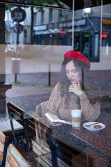 Pretty woman with curly hair in red french beret outside the cafe window reading the book with a cup of latte
