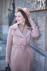 Pretty woman with curly hair in pastel pink coat near stone wall in autumn park holding her hat by left hand