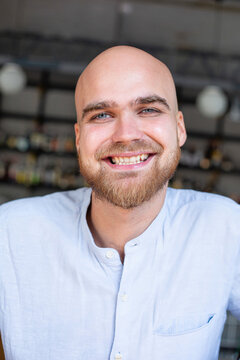 Portrait Of Smiling White European Bald Man With Beard And Grey Eyes In Light Blue Shirt In The Workspace