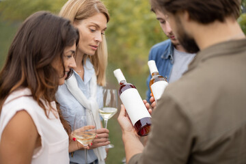 Joyful european friends tasting local wine near vineyards in countryside. Man showing a bottle with blank label to copy paste. Young men and women enjoying time together