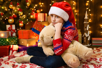 Child girl posing in new year or christmas decoration. Festive lights and lots of gifts, an elegant Christmas tree with toys. The girl is wearing a red sweater and a Santa hat, she playing with bear