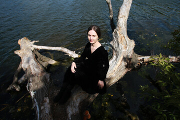 Brunette woman in black dress sitting on a snag near the lake
