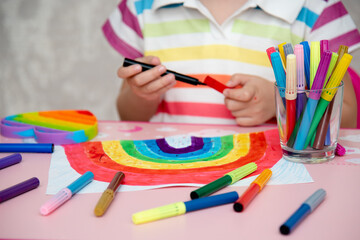 A cute girl in rainbow clothes draws a rainbow with multi-colored markers.