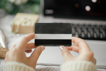 Credit card in hands on background of laptop with black screen on stylish bed with modern christmas ornaments and gift. Christmas sales and shopping online. Space for text. Black friday