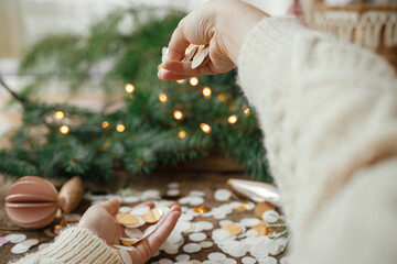 Fototapeta na wymiar Hands in sweater holding golden confetti on background of christmas lights, decorations, baubles and pine branches on rustic wood. Festive mood. Space for text. Happy holidays. Xmas party