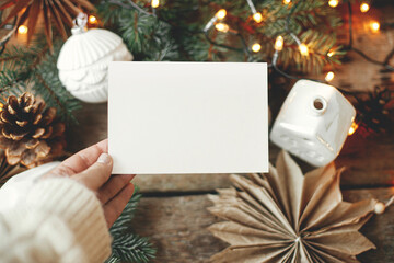 Christmas card mock up. Hand holding empty greeting card on background of christmas stars, baubles, cones and pine branches in lights on rustic wood. Space for text. Seasons greetings template © sonyachny