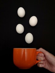 Chicken eggs of white color fly out of an orange cup in hand on a black background