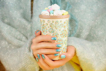 Female hands with blue festive manicure holding a cup of cocoa and marshmallows. Hot cocoa on a snowy day. Christmas holidays.