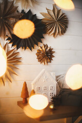 Obraz na płótnie Canvas Stylish christmas little trees and house on background of white wooden wall with big paper stars and christmas golden lights bokeh. Festive decorated scandinavian room. Atmospheric eve