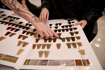 Naklejka premium Close-up of a well-groomed tattooed hairdresser arm pointing to a swatch of straight dyed hair in a hair dye color palette catalog and advising a client on hair coloring in beauty salon