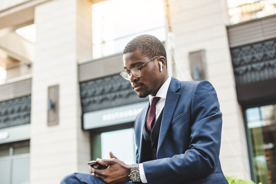 Portrait of black african american businessman in suit with glasses and headphones sits on the background of city buildings outdoors