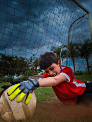 Boy struggles to catch the ball playing football (soccer) as a goalkeeper with dramatic sky in the...